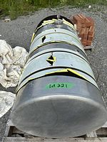 Top-down and side views of the heavily damaged cylindrical metal fuel tank sitting on a wooden pallet outdoors, showing large holes and tears sealed with yellow tape.