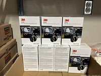 Seven boxed 3M Peltor X2P5E ear protection mufflers stacked on a shelf, showing front of boxes with worker wearing ear protection illustration.
