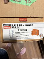 Close-up photo of the box label showing Simpson Strong-Tie LUS28 Hanger details including quantity (50), sizes compatible (2x8 and 2x10), and included screws and nails.