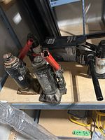 Photo of four used hydraulic bottle jacks with red handles on a shelf, metal construction, black paint with wear marks.
