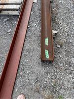 Two steel I-beams lying on gravel ground with pallet to the left and container to the right, both labeled 2x10.
