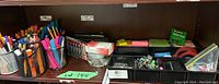 Photo showing assorted colorful pens and markers in metal mesh pen holders alongside tape rolls and binder clips arranged in black plastic bins.