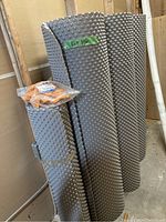 Three rolls of grey dimple board membrane leaning against wall with a sealed bag of orange plastic fasteners on top of the front roll