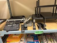 Overview of metal shelves with various office supplies including label maker, staplers, hole punch, and storage trays.