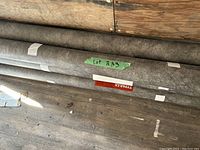 Six grey fabric rolls lying on wooden floor against plywood wall, green tape label visible on one roll