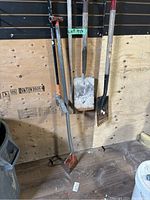 Photo showing all six garden tools hanging on a wood wall, including three shovels, one Fischer weed picker, one garden rake, and one blade tool.