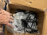 Black metal peg board hooks in a cardboard box, some wrapped in clear plastic bags.