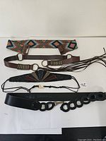 Four belts laid out: top is a bright multi-colored beaded belt with geometric pattern, below is brown leather belt with rings and fringe, then a black vintage beaded stretchy belt with fan design, and bottom a black Danier leather belt with circular linked buckle.