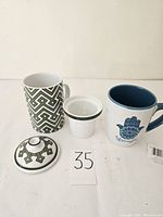 Photo of two mugs: one tea mug with removable strainer and lid featuring a green geometric pattern, and one mug with a blue interior and 'Strength' hand graphic.