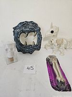 Photo of snarling cat wall hanging, skeletal unicorn figurine, skull bottle stopper in box, and bone drink opener on purple backing