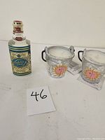 Photo showing one bottle of traditional 4711 cologne alongside two small decorative candles in silver metal buckets with sun faces painted on them.