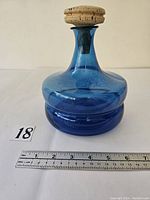Turquoise blue glass pitcher with cork stopper shown with measuring ruler for size reference.
