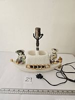 Full view of the vintage lamp showing the boat-shaped ceramic base with two Chinese-style figures, lamp socket in center, gold trim, and attached power cord.
