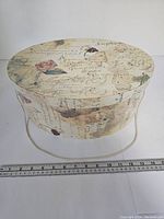 Round decorative box with vintage script and rose design, default closed view with ruler for scale.