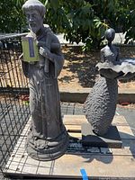 Photo of two garden statues on pallet outdoors: left is a religious figure holding an object with a cross necklace; right is a flower petal style female figure on square base.