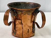 Front view of the antique copper cup showing its wide opening, two large handles, and aged patina on the surface.