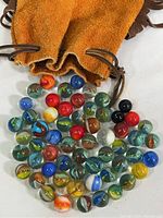 Various vintage glass marbles of multiple colors and sizes spilled from an orange-brown leather pouch with a drawstring closure.