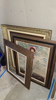 Three vintage framed art pieces stacked leaning against wall - two with artwork, one empty frame