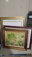Stack of 7 framed art pieces leaning against a wall, showing variety of frame styles and sizes