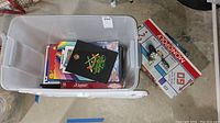 Plastic storage bin containing assorted board games and two books, opened to show contents.