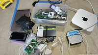 Photo showing various electronics including Apple Mac Mini, tablets, phones, computer memory sticks, and cables all placed on a floor surface.