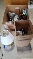 Photo showing two large boxes containing various lighting items such as lamps, light bulbs, and accessories. Visible is a small table lamp with white fabric shade and brass base.