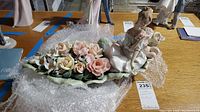 Porcelain figurine on leaf base with many pink and cream flowers, some damaged, placed on wooden table with bubble wrap underneath.