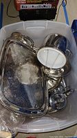 Plastic bin filled with various vintage silverplate servingware items including trays, ice bucket, coffee pot, and other pieces. Some items show wear and damage.