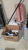 Cardboard box containing various tools and accessories next to rolls of kraft paper and a metallic floor roller on a basement concrete floor.