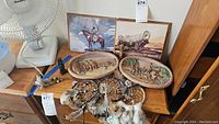 Photo showing two framed western paintings, three oval 3D art plaques, three feathered dream catchers, and a small totem pole figure arranged on a wooden table.