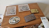 Overview of all five belt buckles on wooden surface including two in plastic cases, one with floral ceramic inlay, gold-tone block letter buckle, and rectangular embossed buckle.