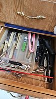 View of wooden drawer with plastic organizer tray containing various kitchen knives with wooden and plastic handles, kitchen tongs, and a manual can opener.