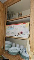 View of wooden cabinet shelf containing boxed Corelle winter-themed beverage set, stemmed glasses and a short tumbler with decorative images at back shelf, and stacked Corelle plates and mugs with green leaf design on bottom shelf.