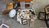 Overview photo showing a pile of assorted collectible spoons, necklaces, pendants, a white display plate with scalloped edges, small boxes containing jewelry, tools, gears, pins, and cases on carpeted floor.
