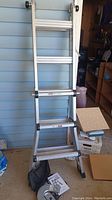 Full view of silver extendable Gorilla Ladder leaning against a blue wall, showing ladder rungs and black locking mechanisms on the sides.