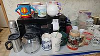 Photo of microwave, coffee mugs, teapots, thermal cups, glass coffee pot, plastic containers and measuring cups arranged on countertop.