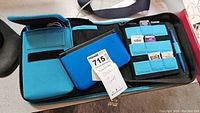 Photo showing black Nintendo 3DS console, blue and black carrying case with elastic slots holding multiple game cartridges, and hanging charm attached to case zipper.