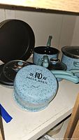 Cookware set showing blue speckled pots and frying pan with green handles and lids, one pot bottom showing Paula Deen logo