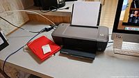 Photo of the HP Deskjet 1000 printer on a desk with paper loaded and a red item nearby.