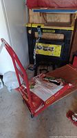 Red metal hand truck with a bag of garden soil and white sack. Black and yellow Storehorse folding saw horses in background, stored in a shed.