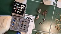 Lot laid out on green tiled surface showing multiple silver jewelry pieces including rings, necklace with heart pendant, hoop earrings with blue beads, and stud earrings with colored hearts in box.