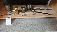 Full view of three sterling silver cups, assorted cutlery including fork, spoon, and large knife with guard on wooden surface.