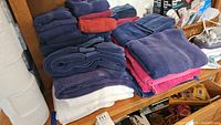 Stack of assorted bath and hand towels in navy blue, red, white, and pink on a wooden shelf.