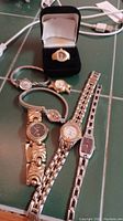 Five wristwatches laid out on green surface, plus marquise-shape watch face ring box