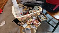 White laundry basket on stool filled with mixed costume jewelry, watches, medallions, key rings