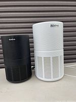 Two Renpho branded air purifiers, one black and one white, shown side by side on a surface with a corrugated background. Both units appear clean and in working condition with visible power cords.