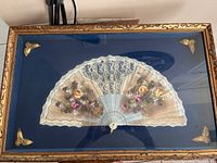 Front view of an antique fan in an ornate gold frame with blue fabric background. Close-up to show lace and floral details on the fan.