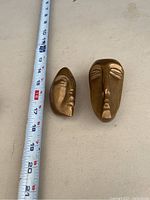 Two brass face sculptures shown next to measuring tape for scale, each about 12 cm tall, made of solid brass with stylized abstract human face designs.