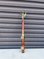 Photo of a single elephant ankush with wooden and brass details, set against corrugated metal background.