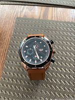 LIGE wristwatch with round black dial, rose gold-tone case, and brown leather strap shown on a textured woven mat surface.